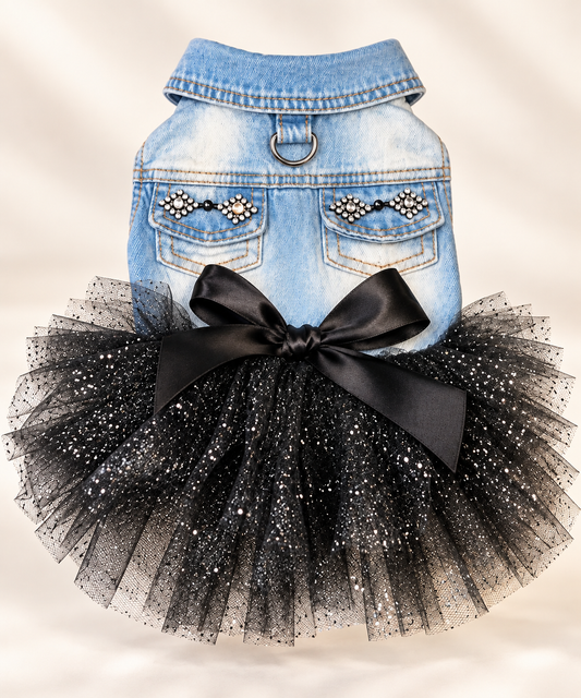 A pet outfit featuring a light blue denim vest with decorative rhinestone pocket details and a voluminous black tulle tutu skirt with a large black satin bow, shown against a soft beige background.